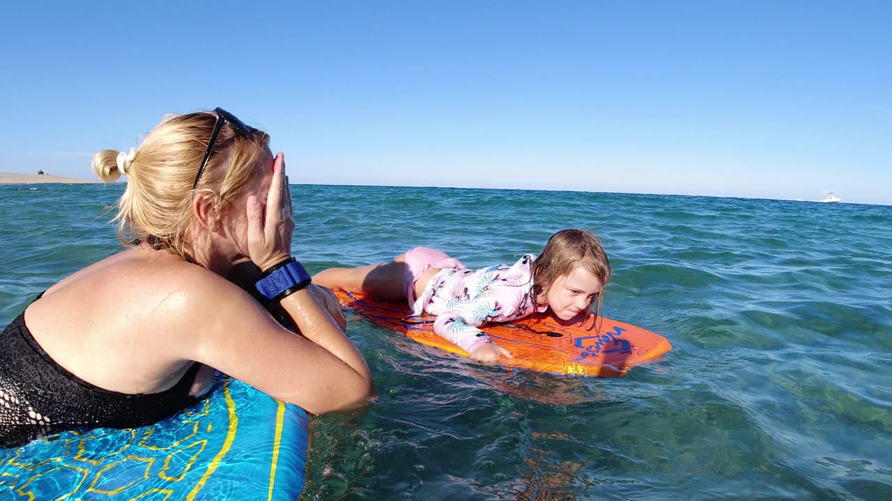 Woman on a blue float watching a girl on an orange board in the ocean.