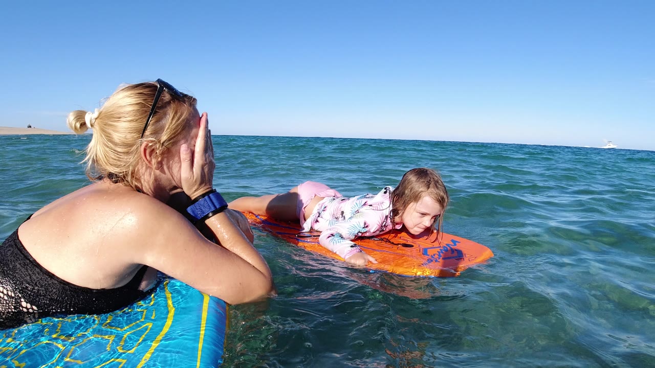 At 0:01 in Los Cabos, Mexico, the woman keeps her hand near her face while the girl looks down from the orange board as small waves pass between them.