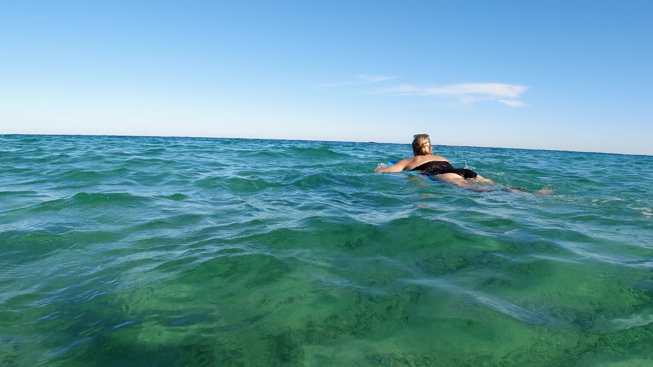 Water-level view in Los Cabos, Mexico of a woman in a black swimsuit floating away from camera on a small board, with green-blue waves in the foreground and a clear sky above.