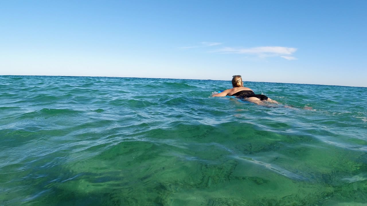 A second water-level frame shows the woman still facing away on the small board, with a slightly higher foreground wave and the bright Los Cabos sky taking the upper half.