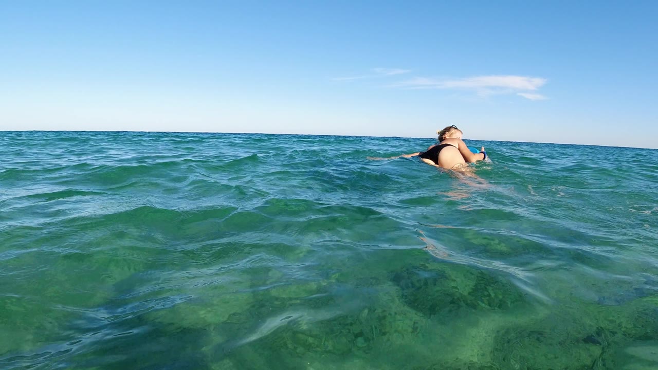The woman turns partly side-on while holding a colorful board in Los Cabos, Mexico, with turquoise water across the foreground and a clean horizon under blue sky.