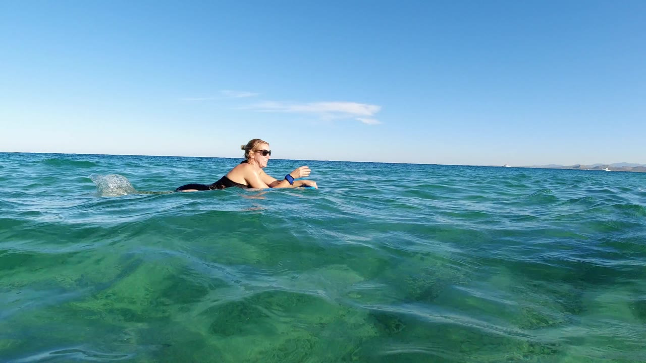 Woman in sunglasses lying on a surfboard in turquoise ocean water.