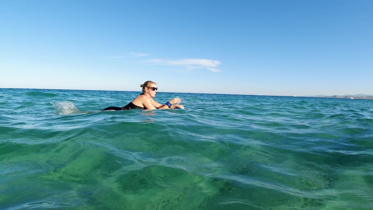 At 0:01, the woman remains centered on her surfboard in Los Cabos water, with a small splash near her legs and the horizon running behind her.