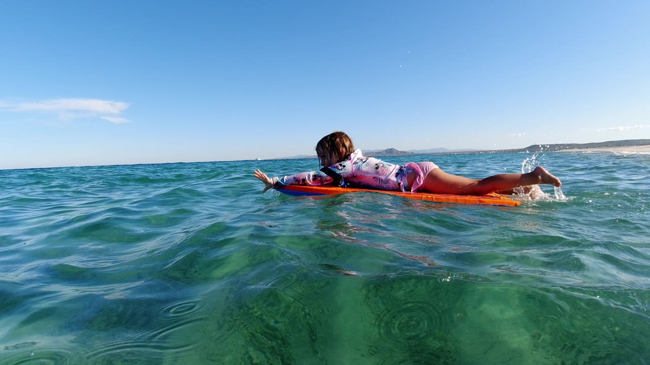 At 0:01, the girl remains stretched across the orange bodyboard as rippled green-blue water fills the foreground and a pale shoreline sits in the distance.