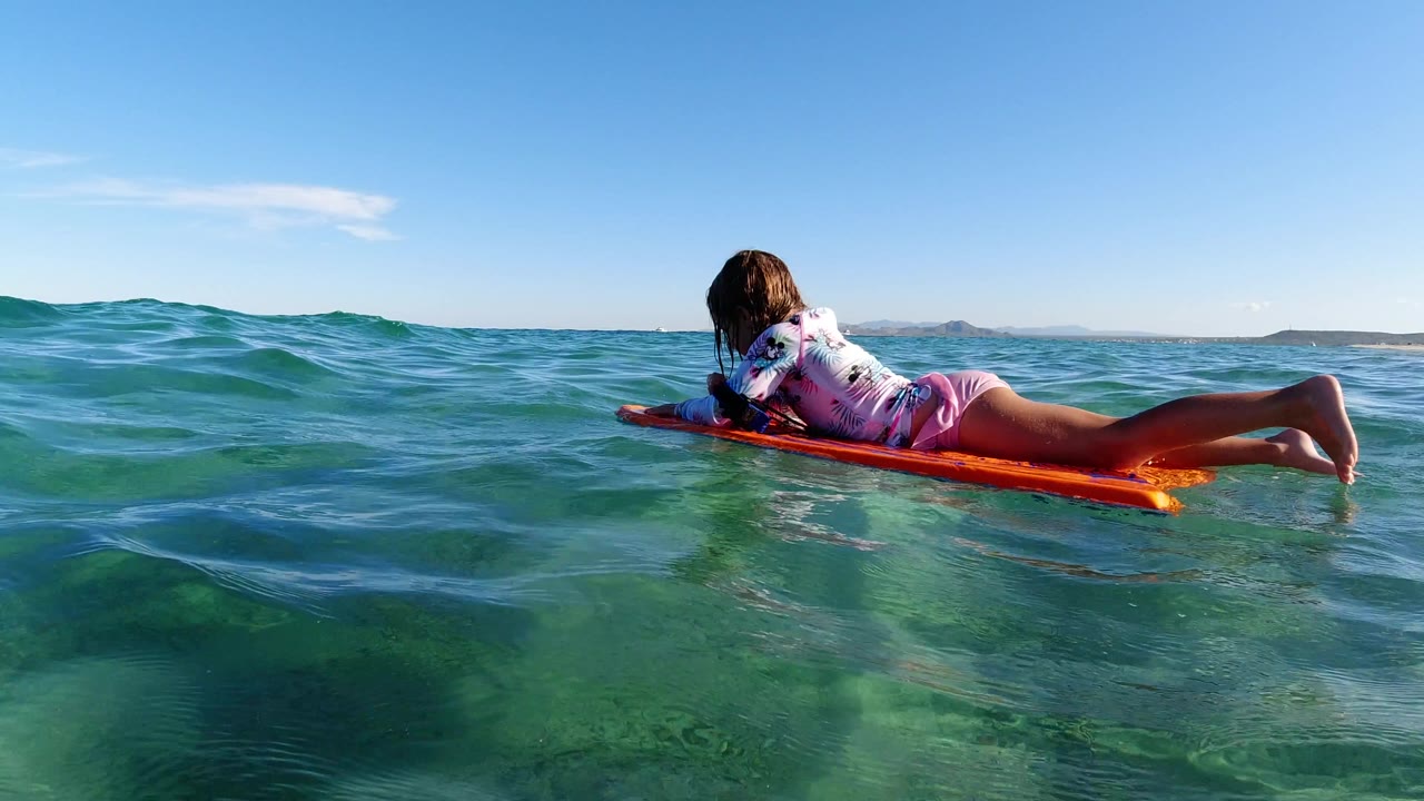 At 0:00, a young girl lies belly-down on an orange board in the clear blue-green ocean off Los Cabos, Mexico, with her body angled away from the camera and the horizon behind her.