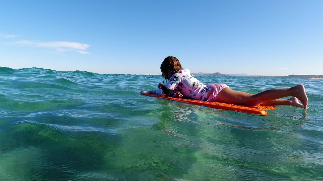 At 0:01, the girl remains stretched across the orange board as a low swell rises in front of her in Los Cabos, Mexico, with bright sky and distant land on the horizon.