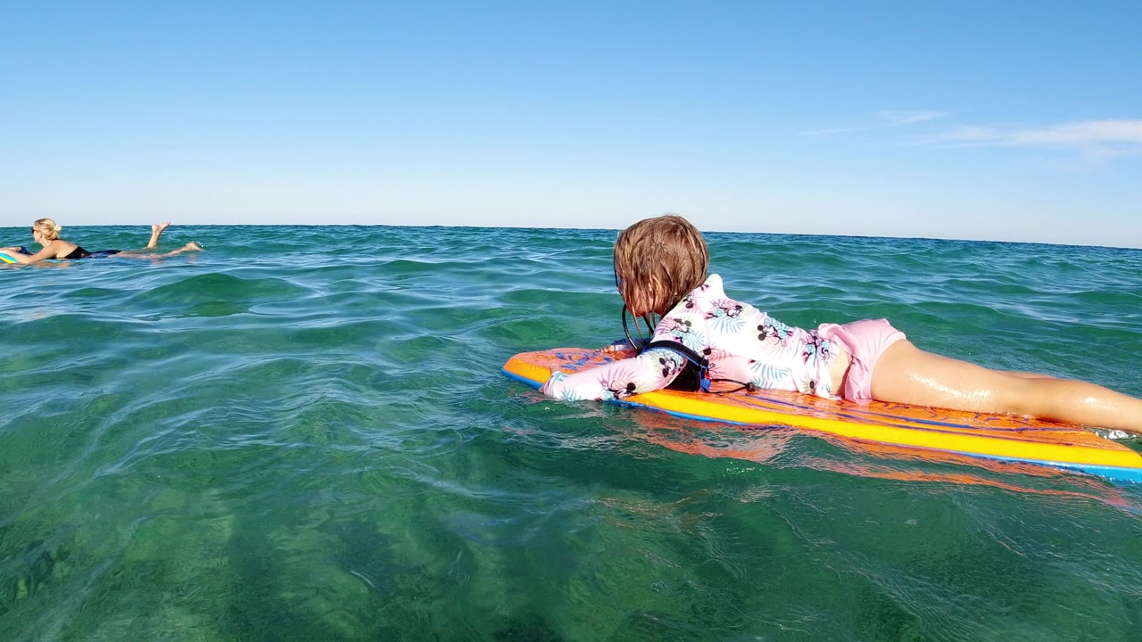 At 0:00, a young girl lies on an orange and yellow surfboard in Los Cabos ocean water while an adult swimmer appears near the left edge.