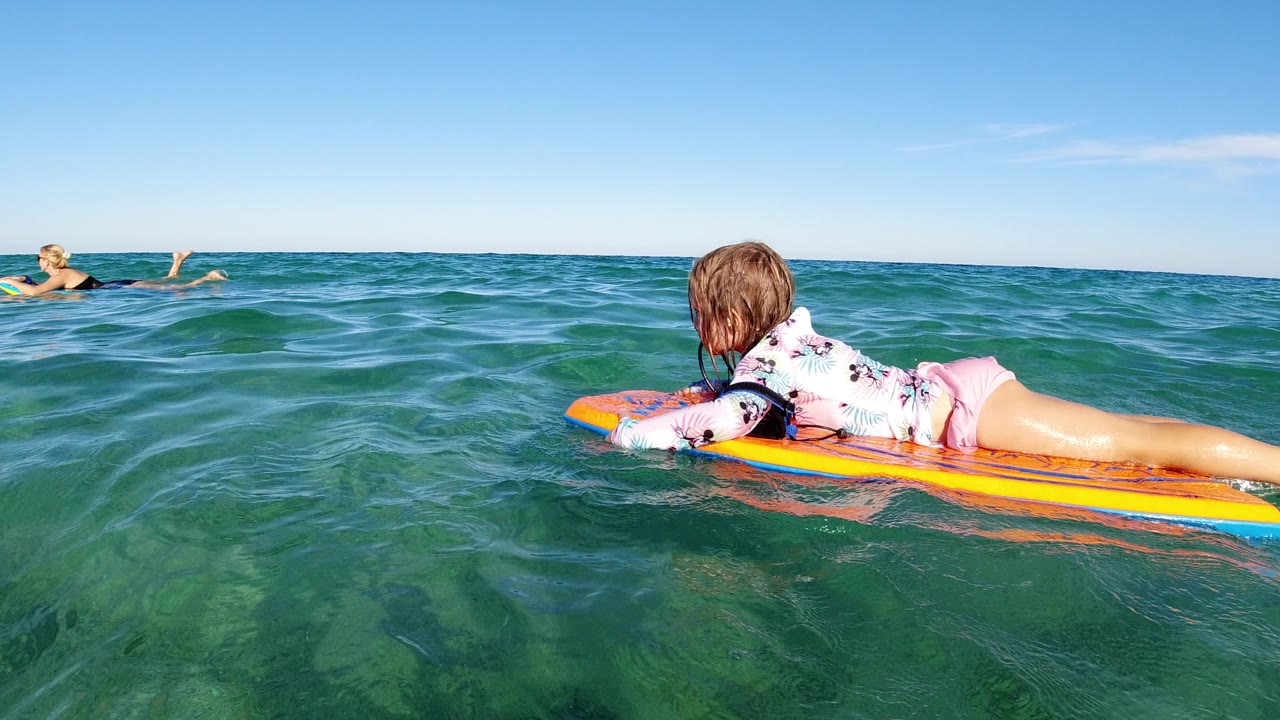 At 0:01, the child stays low on the surfboard as small waves lift between her and an adult swimmer in the Los Cabos water.