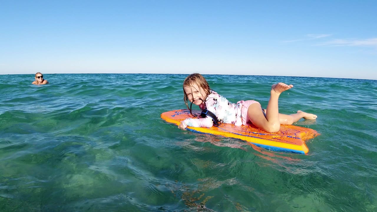 A child rides an orange boogie board near the camera in Los Cabos, Mexico, while another child floats farther back in the open ocean.