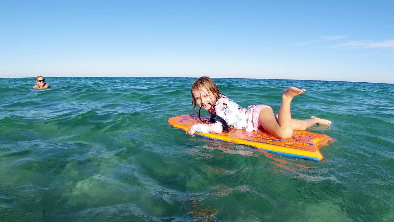 The child smiles toward the camera from an orange boogie board as green ocean water rises around the board in Los Cabos, Mexico.