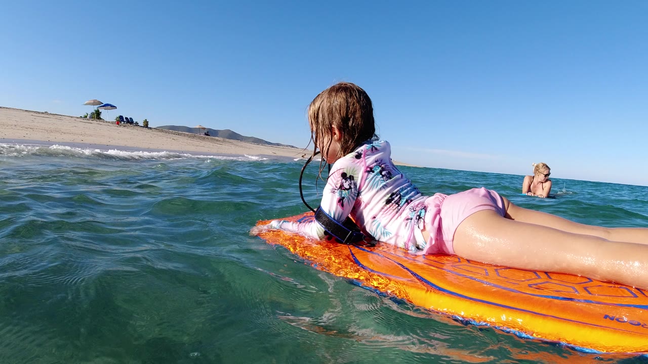 Girl lying on an orange bodyboard in ocean water near a sandy beach.