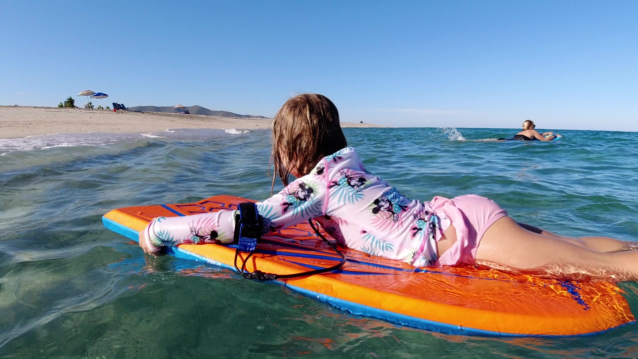 At 0:00 in Los Cabos, Mexico, a young girl lies face down on an orange float board as small waves roll toward the sandy beach and a swimmer appears in the distance.