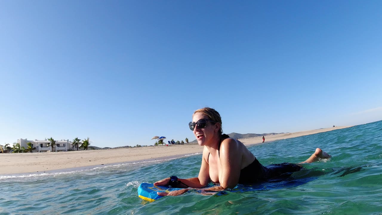 At 0:00 in Los Cabos, Mexico, a woman in sunglasses leans on a blue bodyboard in green-blue surf, with sand, low buildings, and a clear sky behind her.