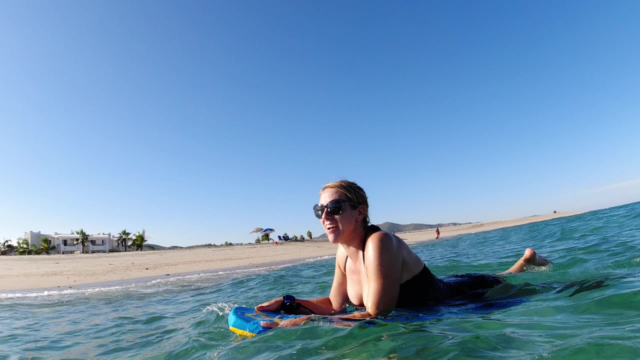At 0:01 in Los Cabos, Mexico, the woman stays low on the bodyboard while small ripples cross the foreground and the sandy beach remains visible on the left horizon.