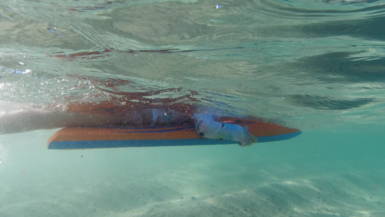 At 0:01, the child stays close to the orange and blue float while sunlight ripples across the shallow water and sandy bottom in Los Cabos, Mexico.