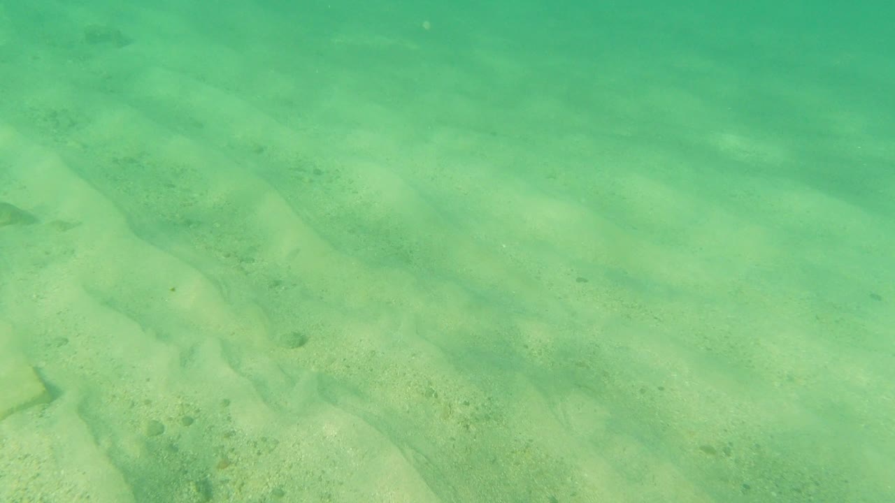 Transparent Los Cabos shallows show pale sand, faint ripples, and diagonal darker current marks, with no visible people.