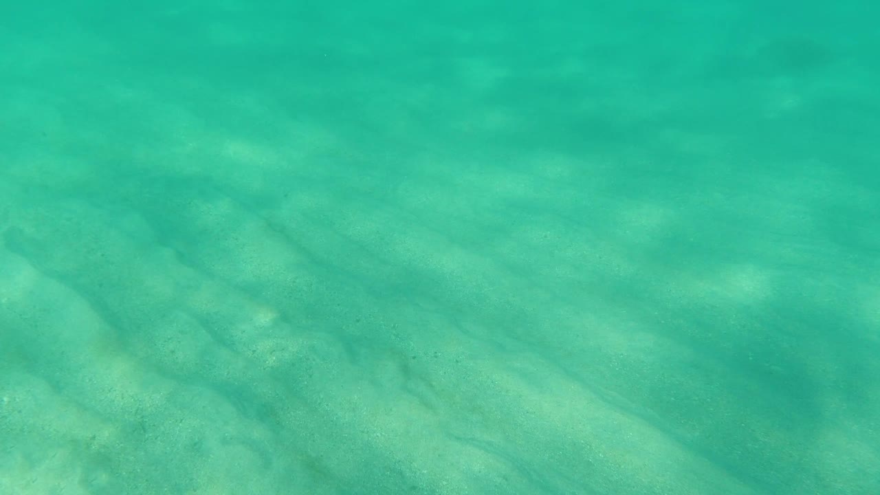 Blue-green water lies over a sandy seabed in Los Cabos, Mexico, with faint ripple shadows and no visible people.