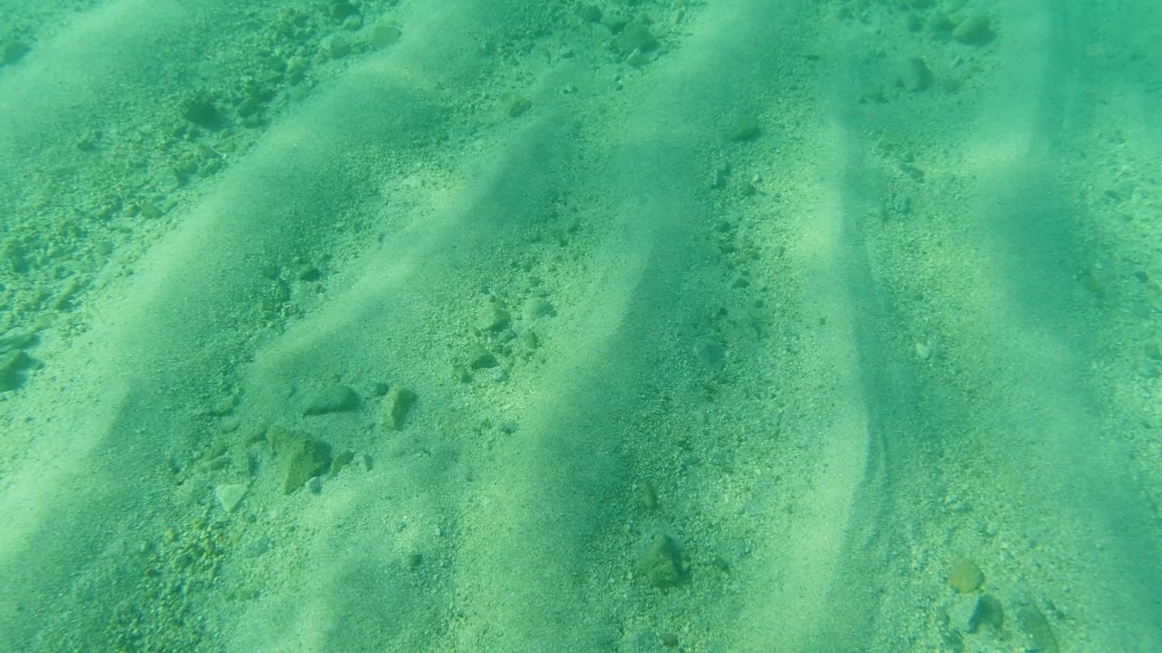 A downward view in Los Cabos, Mexico shows pale sand ridges under green-blue water with scattered pebbles and no visible people.