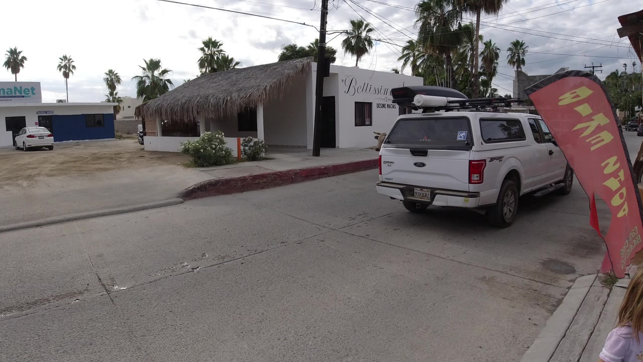 At 0:00 in Los Cabos, a street-level view shows a white pickup beside a red sidewalk flag, storefronts, palms, and a cow just visible near the right edge, with no clearly visible people.