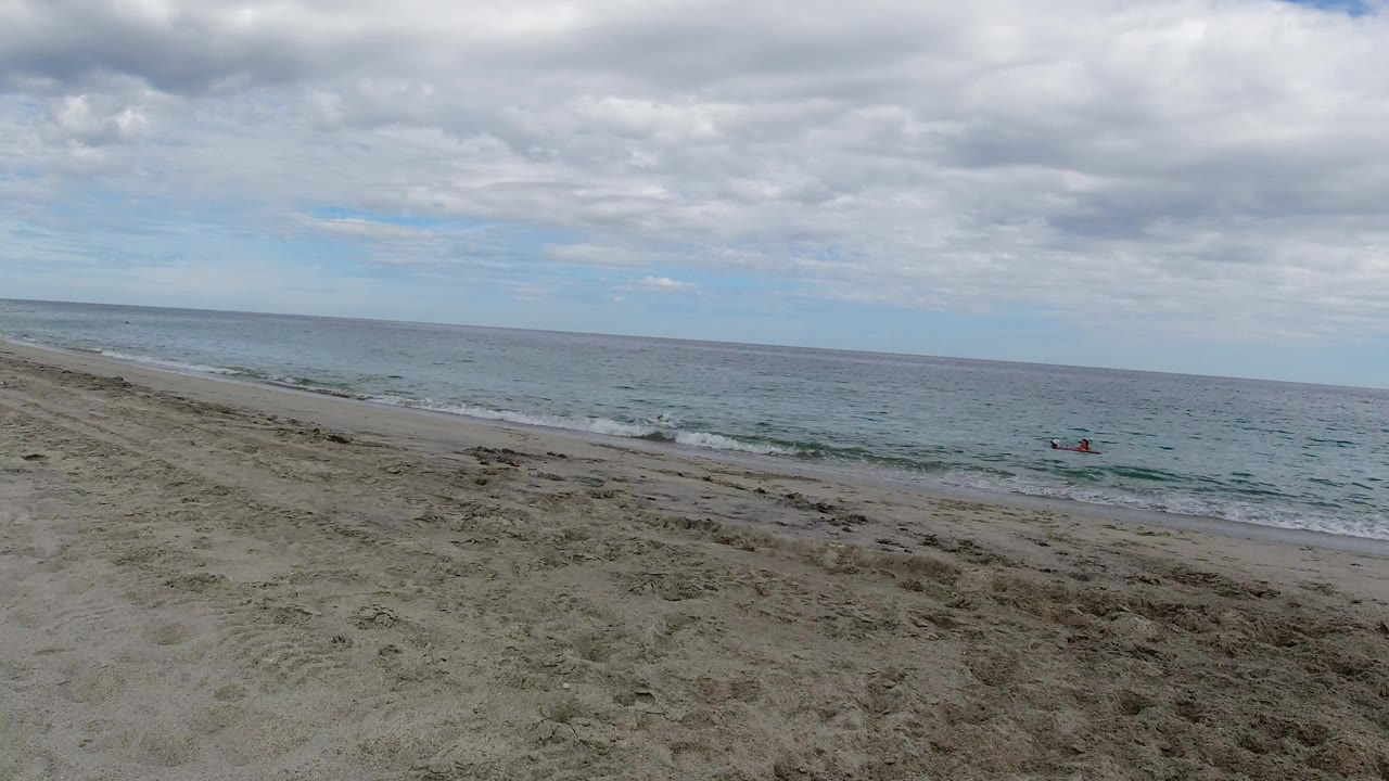 Wide Los Cabos beach view with raked sand in the foreground, low waves near shore, distant red craft on the water, cloudy daylight, and no visible people.