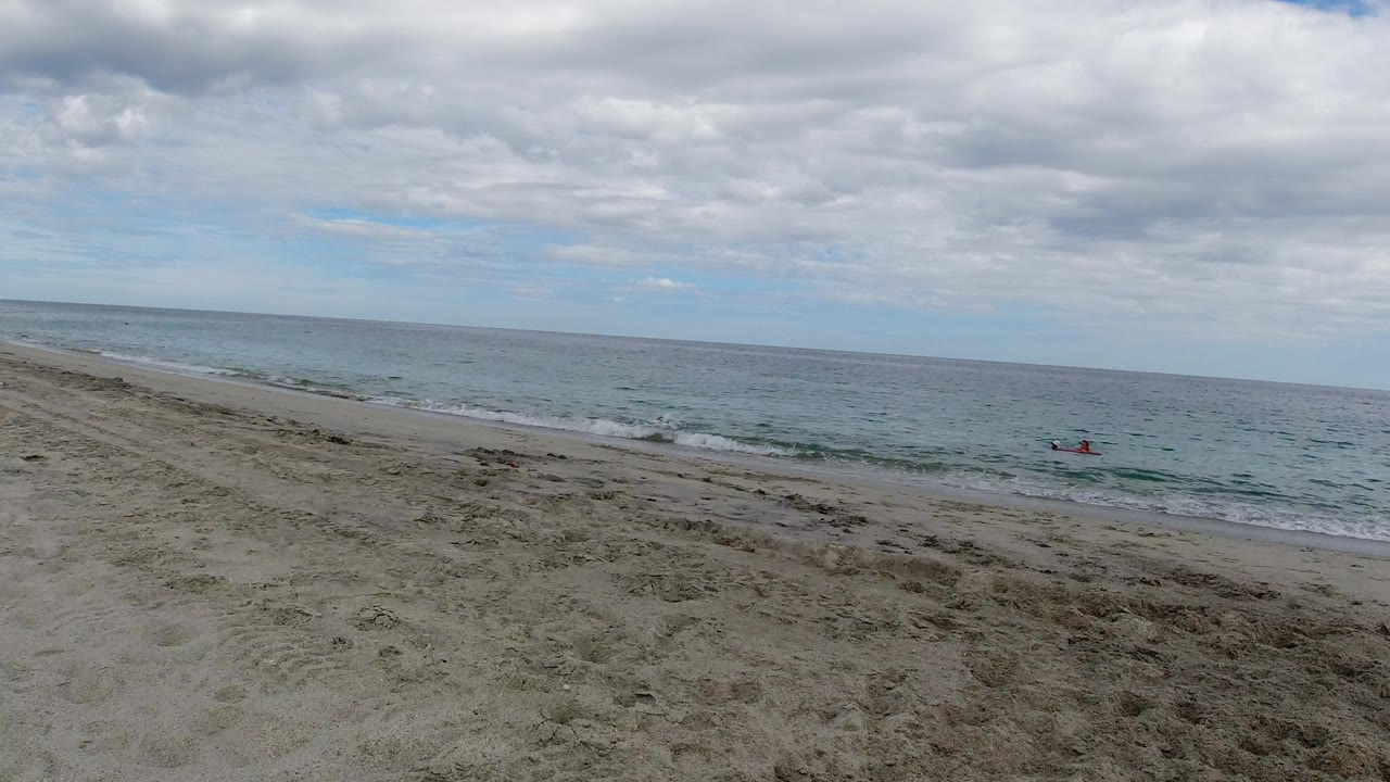 Los Cabos shoreline seen from above the sand, with gentle surf, a cloudy sky, and a small red craft farther out on the water with no visible people.
