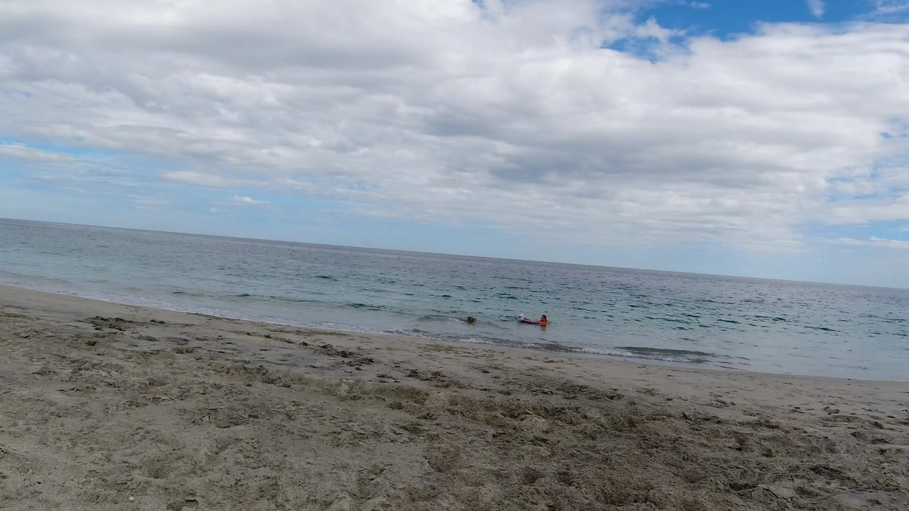 Wide view of a Los Cabos sandy shoreline with low surf, open ocean, heavy cloud cover, and a small distant figure near the water.