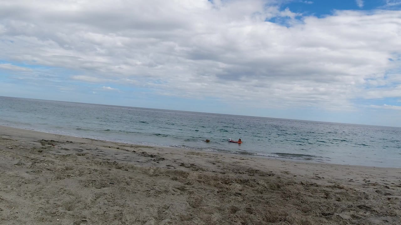 Los Cabos beach view with shallow waves moving toward the sand, two tiny distant people near the surf, and a broad cloud bank over the horizon.
