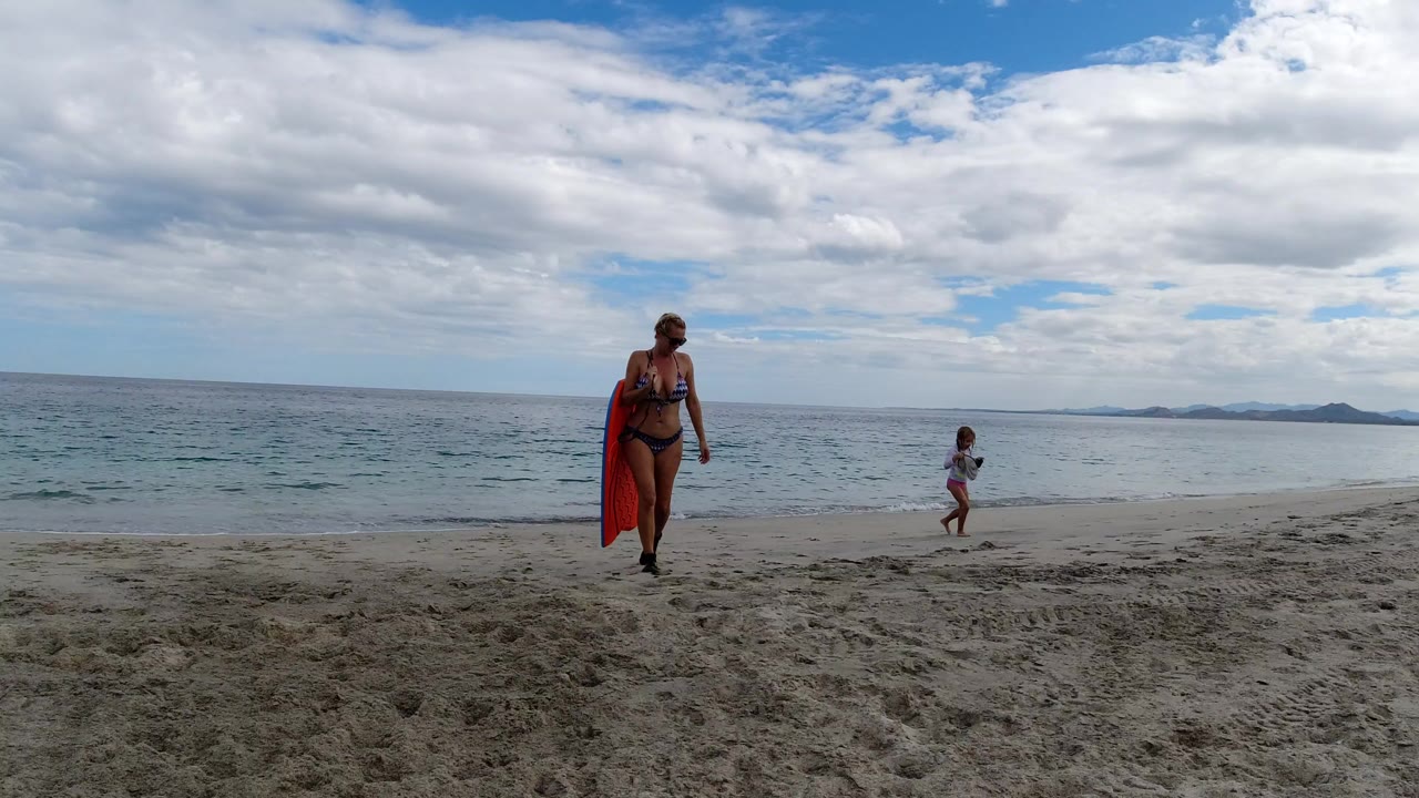 Woman with red board walks on sandy beach as a child stands near the water.