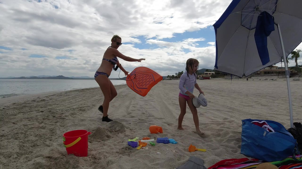 Mother and daughter playing with beach toys under an umbrella on sand.