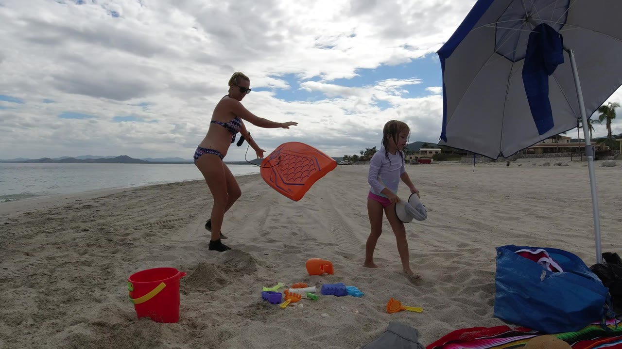 The woman reaches toward the orange mat as the girl stands on the sand near toys, with the umbrella pole, blue bag, calm water, and distant shoreline behind them in Los Cabos.