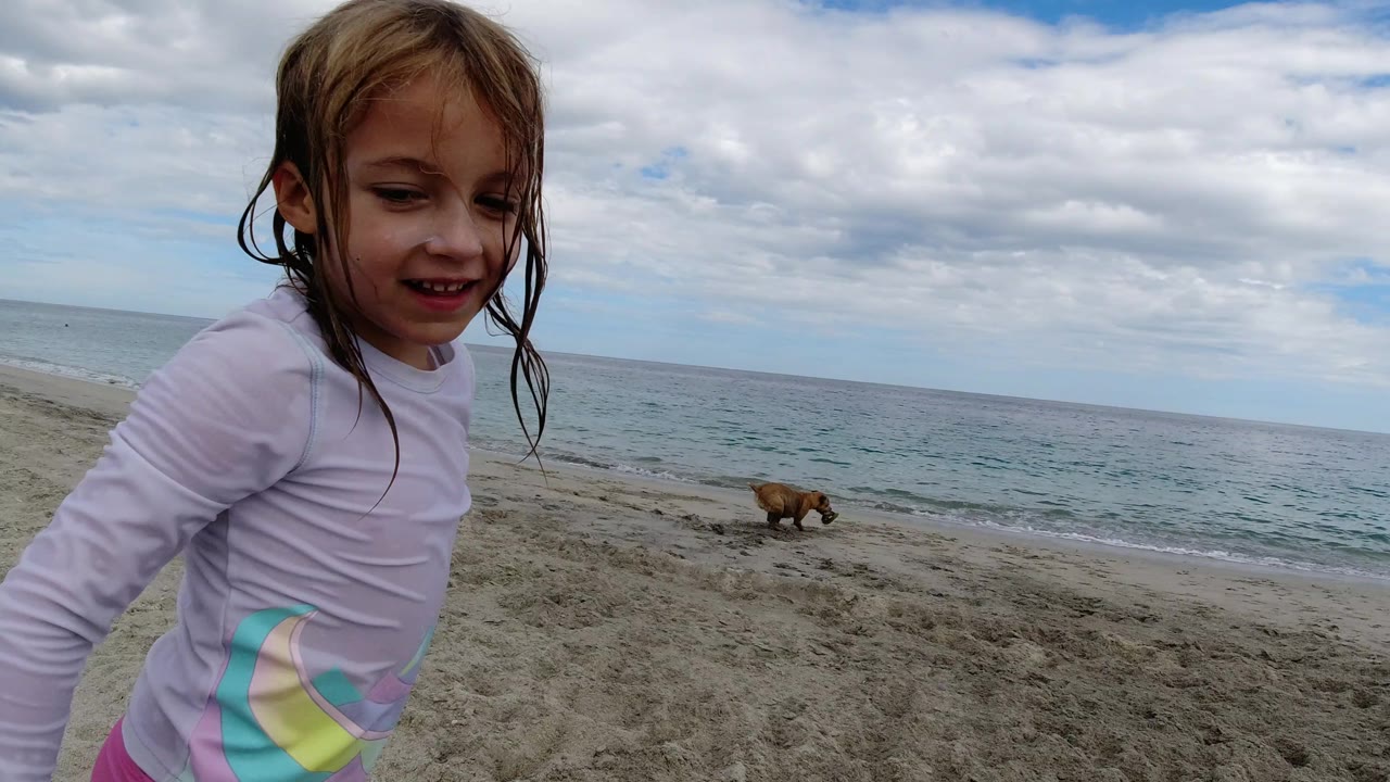 The young girl remains near the left side of the frame on a Los Cabos beach, with the dog farther back on the sand and low waves under a clouded sky.