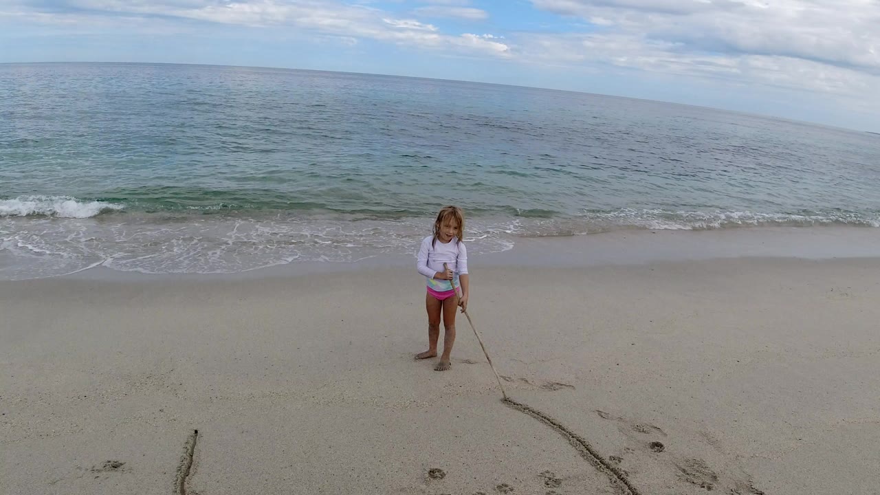Young girl dragging a stick across wet sand near the ocean.