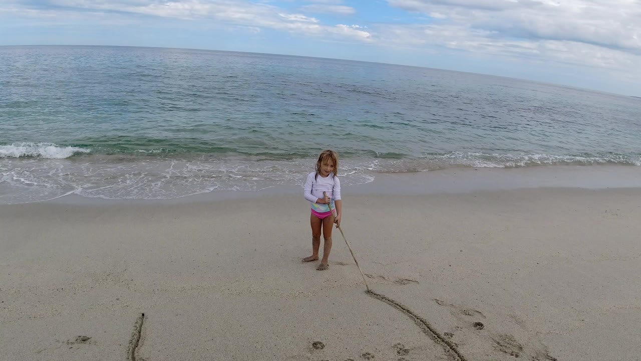 The girl remains near the center-left of the Los Cabos shoreline, holding a stick that trails a dark line across the sand toward the foreground.
