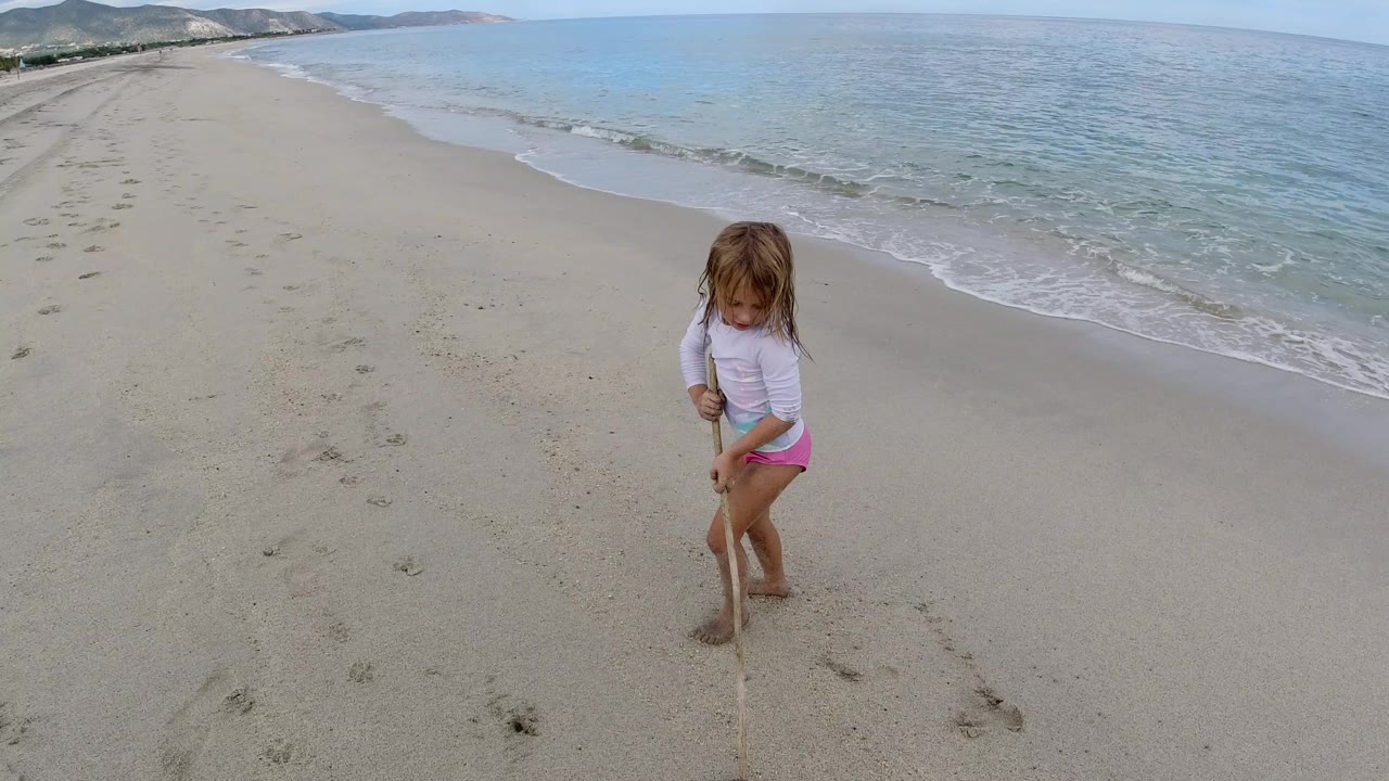 The girl looks down at the sand while gripping the stick on a quiet Los Cabos shoreline, with small waves breaking to her right and footprints crossing the beach.