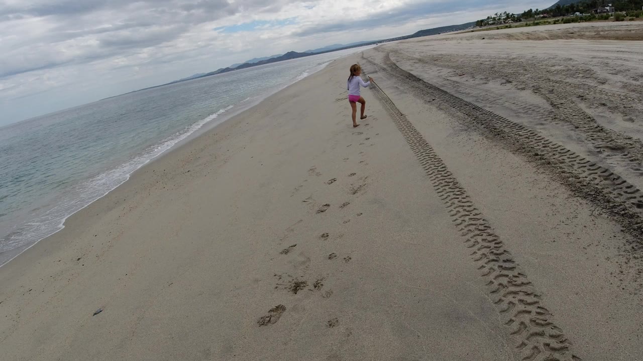 The child moves farther down the Los Cabos shoreline, centered between the waterline and a dark tire track running through the pale sand under broken cloud cover.