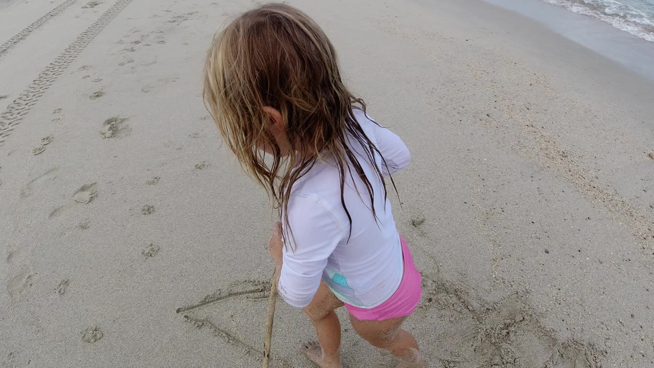 A young girl in a white swim shirt and pink bottoms stands on Los Cabos sand holding a stick near faint surf and beach tracks.