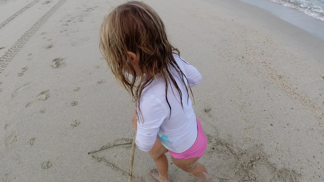 The girl remains near the lower left of the Los Cabos beach frame, with wet hair, a wooden stick, footprints, tire marks, and surf at the edge.