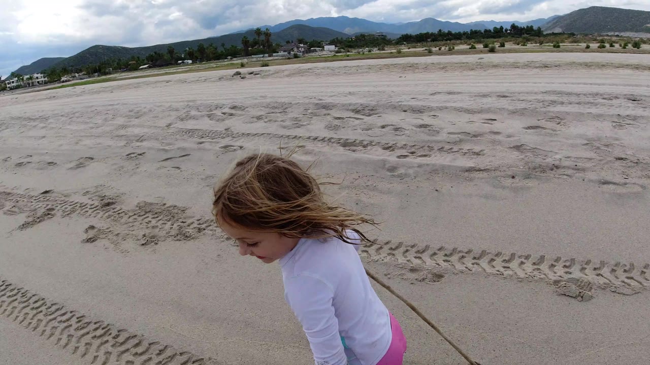 The young girl remains near the foreground edge on the Los Cabos sand as track marks lead across the beach toward distant houses and low hills under broken cloud cover.