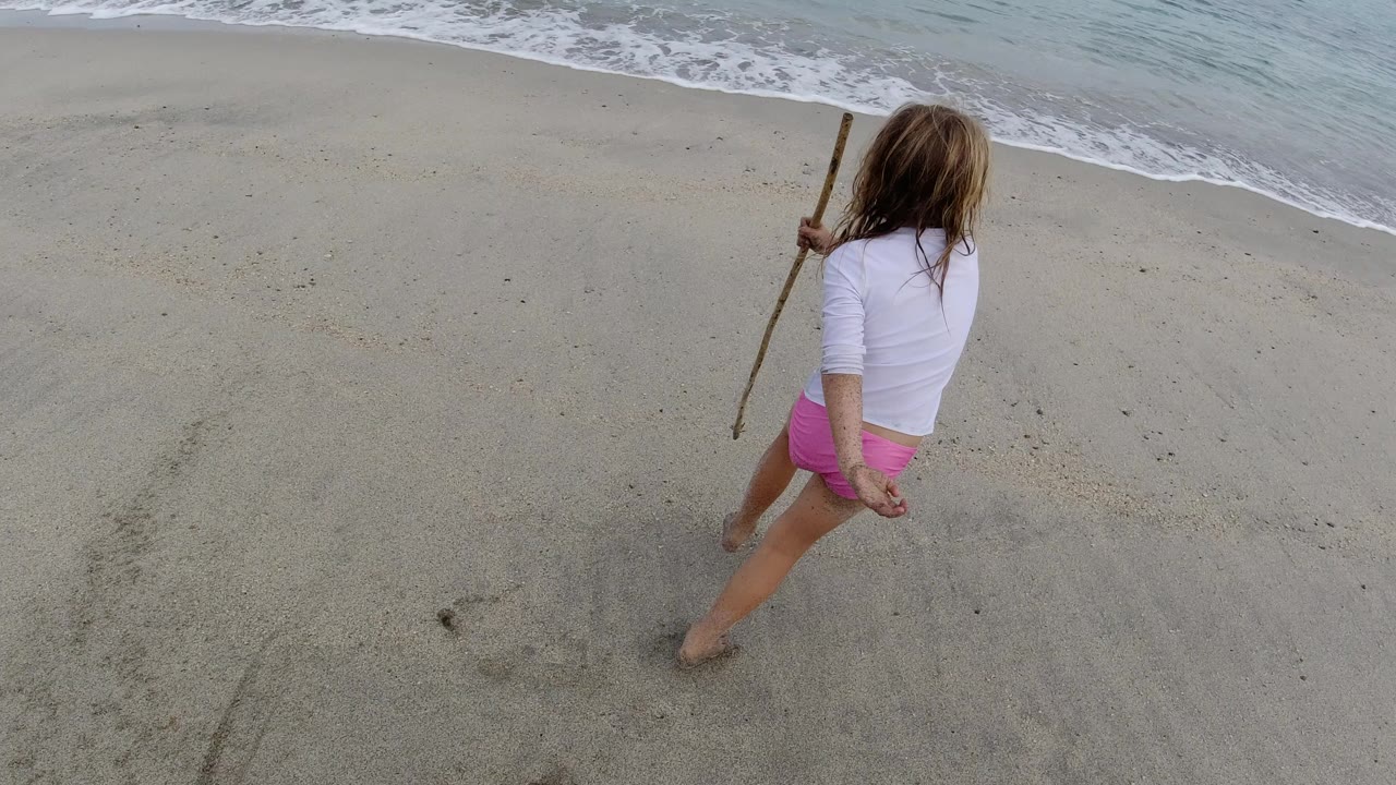A young girl in a white top and pink shorts moves along the Los Cabos beach near the surf, holding a long stick as waves roll in beside her.