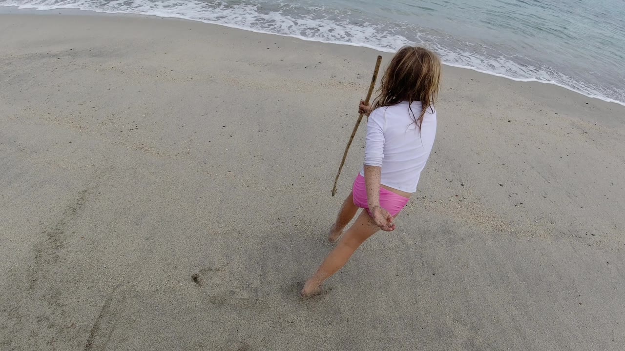 The girl continues moving down the Los Cabos shoreline with her back partly to camera, a stick in one hand and foamy water curving across the sand.