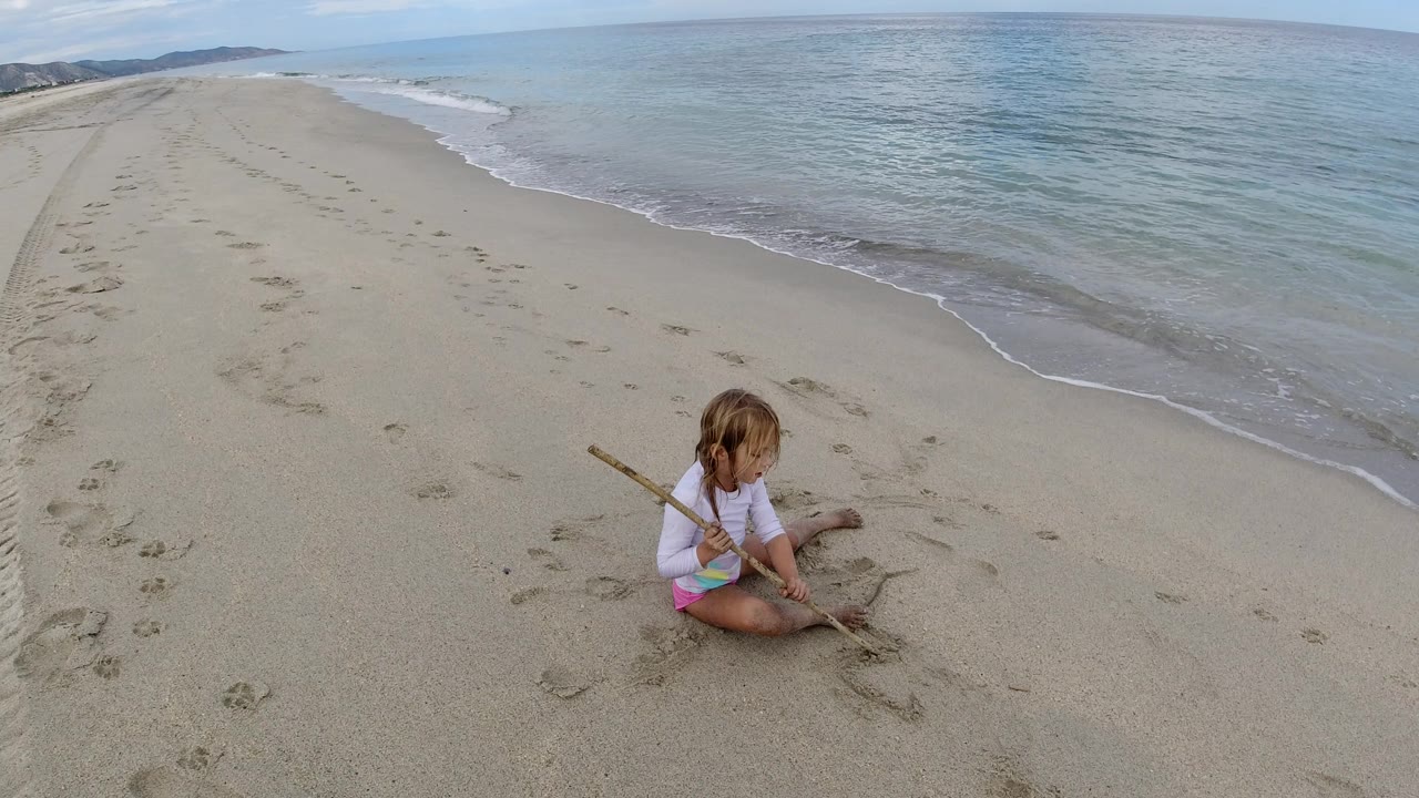 The child remains seated on the Los Cabos beach, angled toward the sand while a thin line of surf curves across the background.