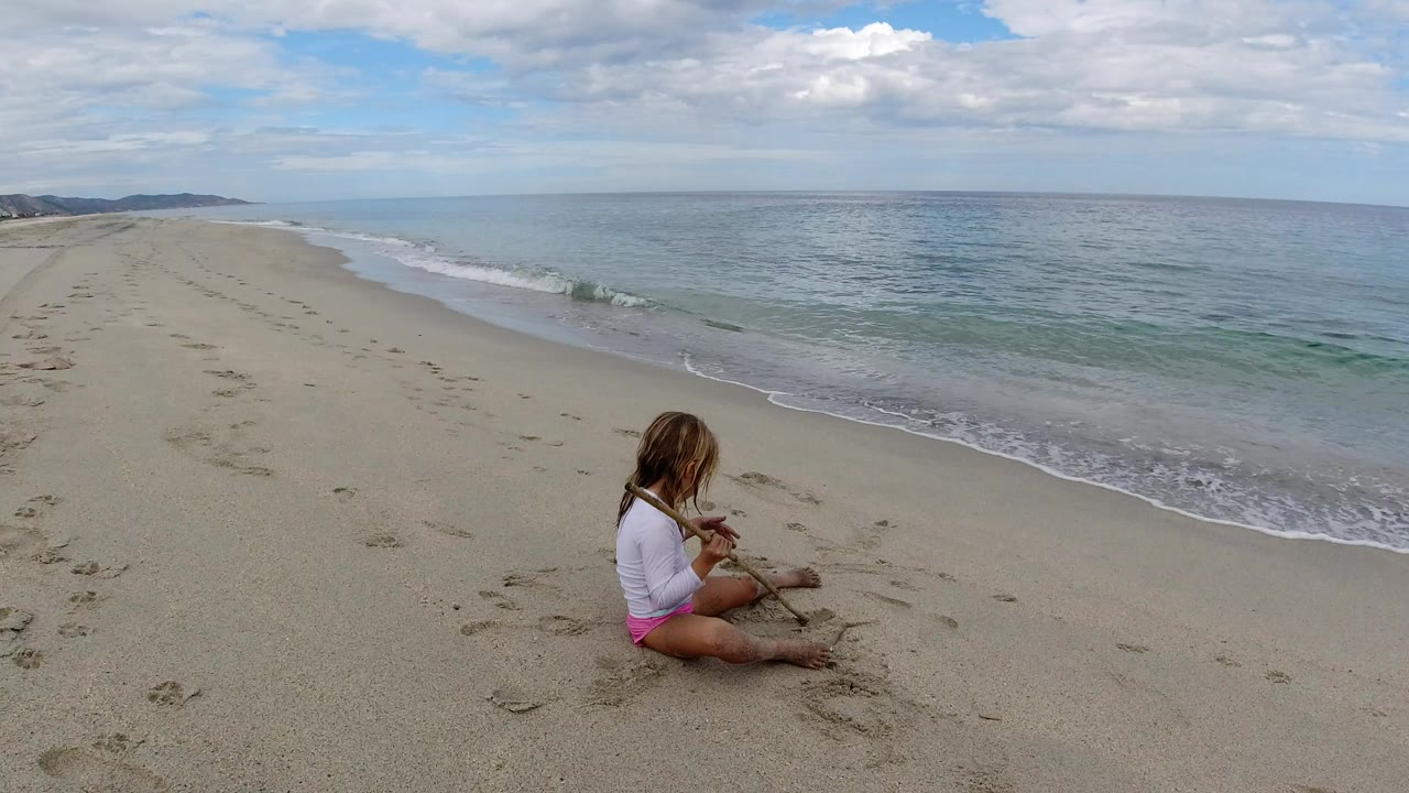 Young girl sitting on sand near the ocean with a stick.