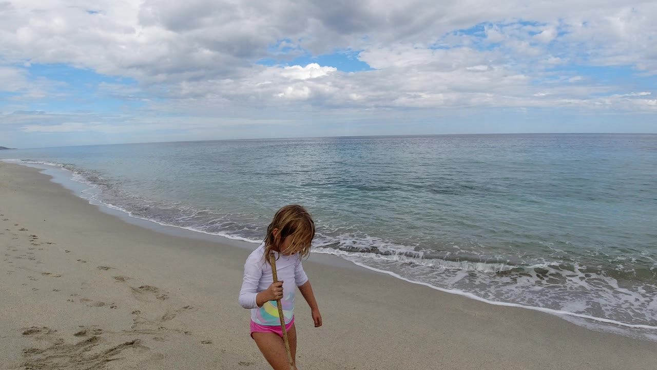The child continues along the Los Cabos beach with the stick held upright while the waterline curves across the sand behind her.
