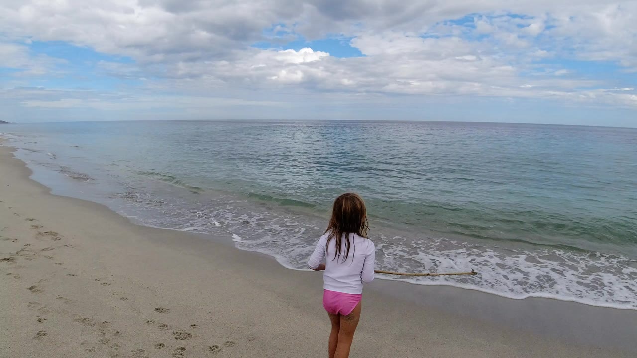 Child standing on a sandy beach facing ocean waves.