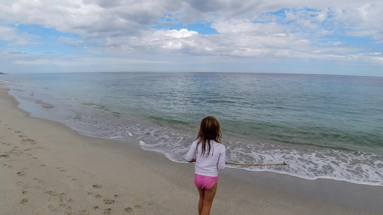 The child remains centered near the lower frame in Los Cabos, Mexico, looking toward a thin line of foam as the ocean rolls onto the pale sand.