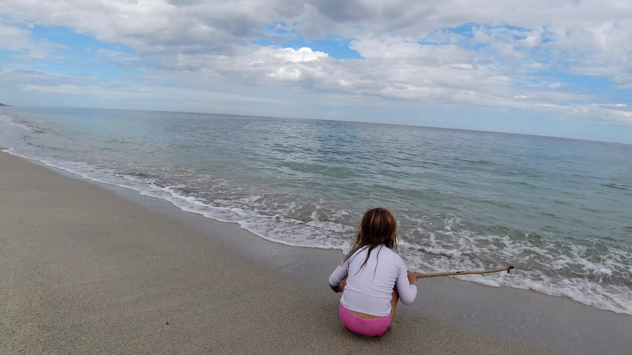 The girl remains seated on the Los Cabos beach, holding the small rod low as a thin wave line reaches the wet sand ahead of her.