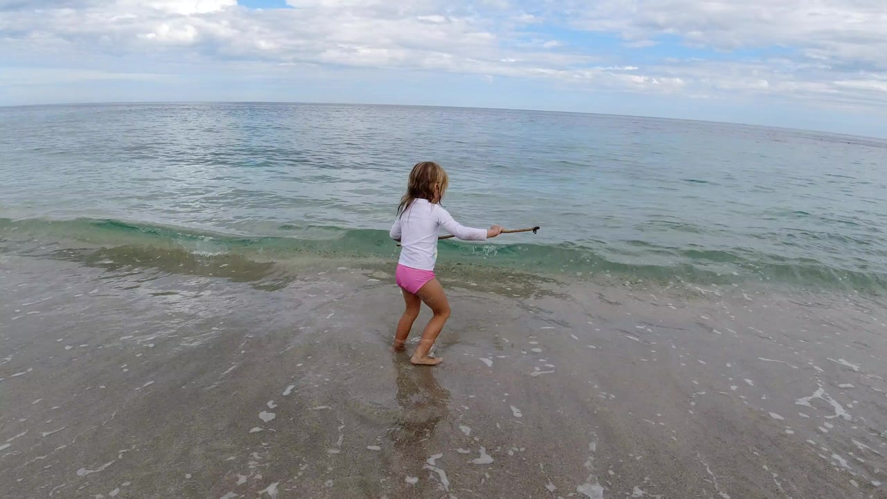 Young girl stands near the waterline holding a stick on a beach.
