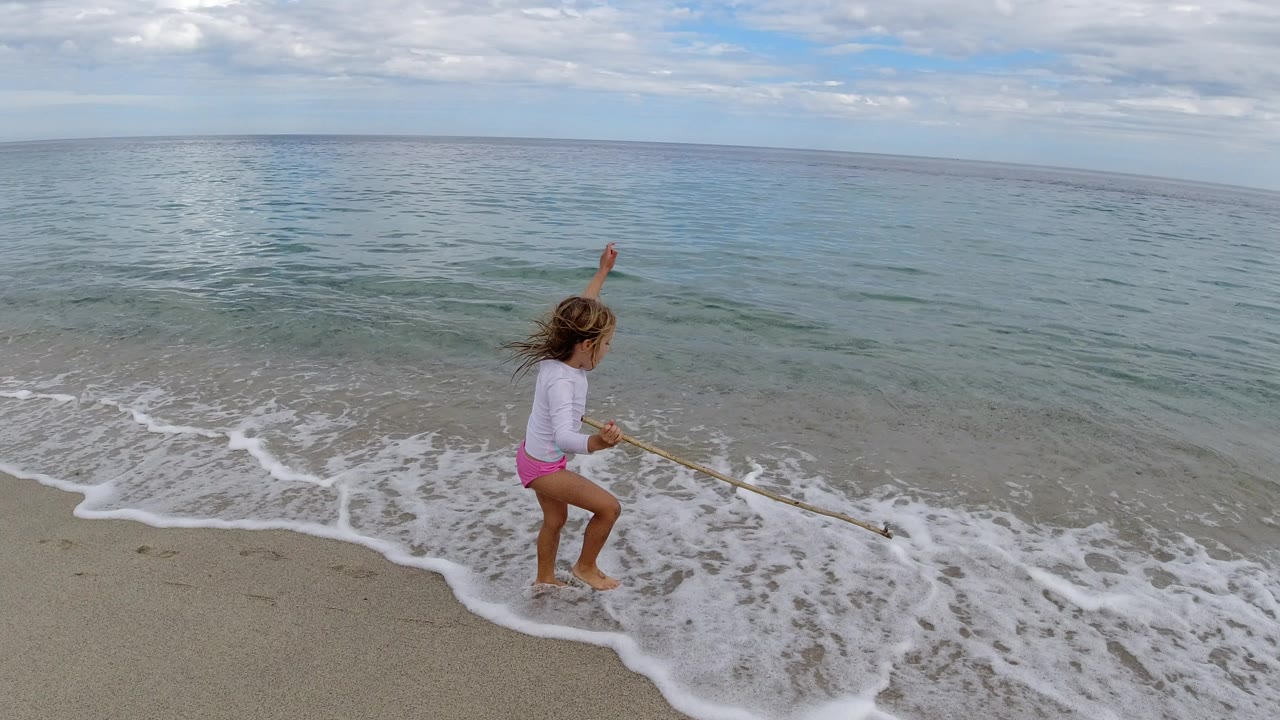 A blonde child in swimwear stands on a Los Cabos beach holding a long piece of wood as a small wave washes onto the sand.