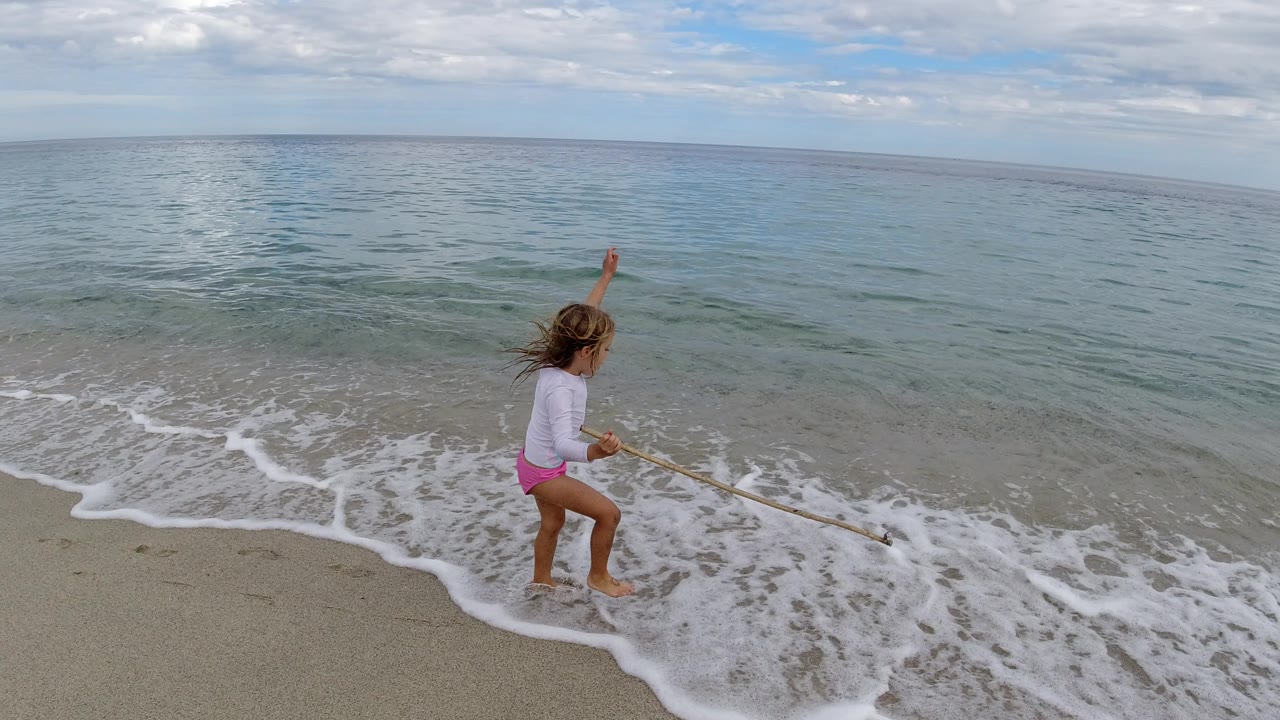 The child lifts one arm while standing beside the incoming Los Cabos surf, with the long wood extending toward the water.