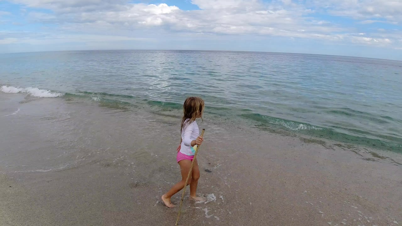 A young girl walks left to right along the wet sand in Los Cabos, Mexico, holding a small stick as the ocean fills the background under broken clouds.